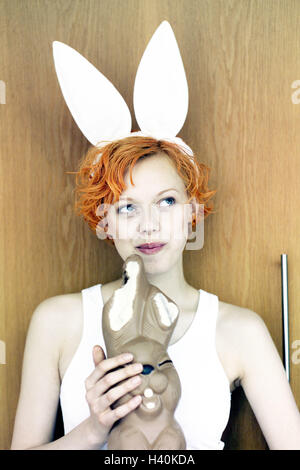 Woman, young, hare's ears, chocolate hare, eat, portrait, young persons, teenagers, short hair hairstyle, flighty, orange, colour hair, expression, smile, smile, happy, view side view, consider, brood, chocolate, Easter bunny, Easter, Easter feast, nibble Stock Photo