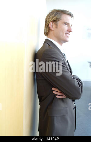 Man, young, contently, half portrait, side view, business, office, businessman, Manager, office worker, blond, blue eyes, suit, stand, lean, lean, wall, smile, calmly, self-confidently, proudly, certainly about victory, successfully, ambition, success, ca Stock Photo