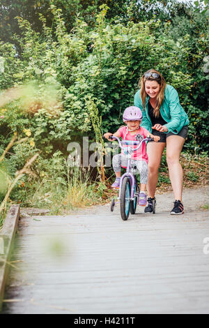 Mother watching daughter riding bicycle with training wheels Stock Photo