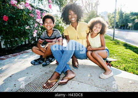 Smiling mother and children sitting on skateboard on sidewalk Stock Photo