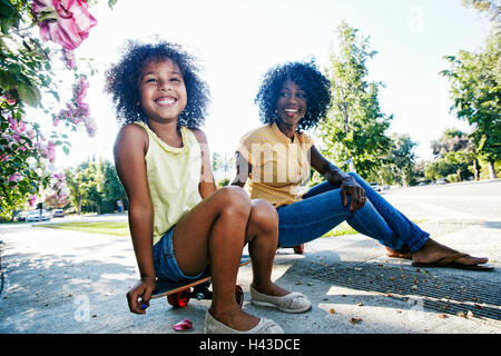 Mother and daughter sitting on skateboard on sidewalk Stock Photo
