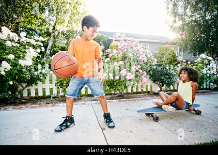 Mixed Race girl on skateboard watching brother dribbling basketball Stock Photo