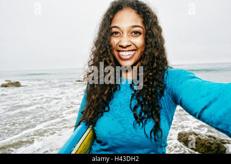 Smiling Mixed Race woman holding surfboard posing for selfie at beach Stock Photo