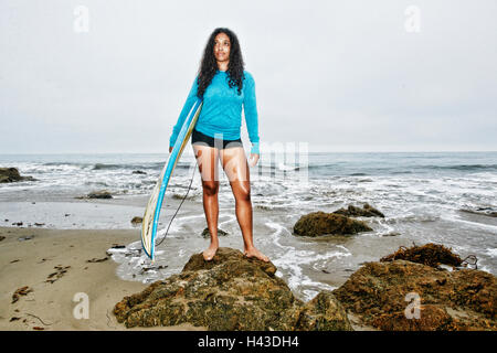 Serious Mixed Race woman holding surfboards on rocks at beach Stock Photo