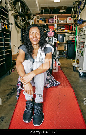 Smiling Mixed Race woman sitting on repair stand in garage Stock Photo