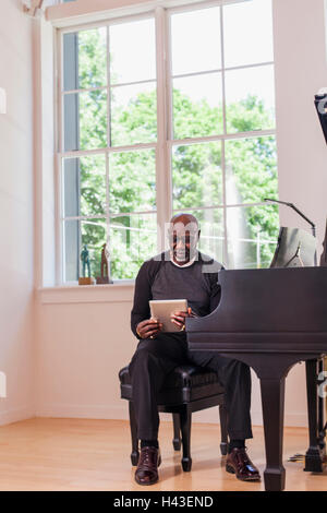 Man sitting at piano using digital tablet near window Stock Photo