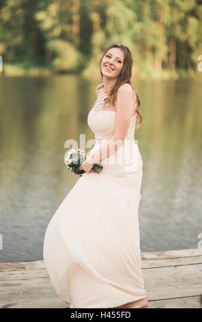 Gorgeous bride in elegant dress holding bouquet posing near forest and lake Stock Photo