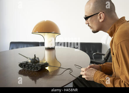 Sits man, table, tanks,,  , plays, profile,  broached,  20-30 years, glasses, glasses bearers, beard, Glatze, lamp, sofa, toy tanks, concentration, fascination, "game child", toy, remote control, war, war game, leisure time, distraction, strategy, remote Stock Photo