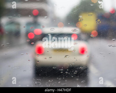 A windscreen with rain drops and light bokeh, whilst driving in the rain. a white, cream car can be seen on the road in front. Stock Photo