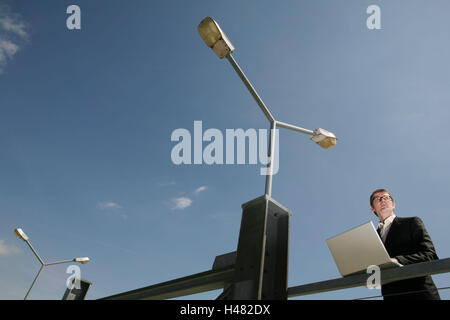 Businessman, seriously, laptop, half portrait, bridge, lanterns, business, person, man, glasses, suit, stand, work, communicate thoughtful, view, foresight, data entry, data transmission, e-mail, Internet, Internet connection, communication, WLAN, without cable, notebook computer, computer, portable, mobility, flexibility, street lamps, outside, Stock Photo