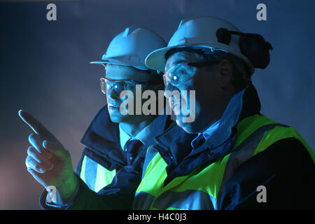 2 senior engineers (one younger, one older) discuss work while pointing. Wearing hard hats, high-viz vests and safety glasses Stock Photo