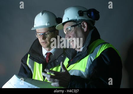 2 senior engineers discuss a drawing in a dark environment. Wearing hard hats, high vis jackets and safety glasses Stock Photo