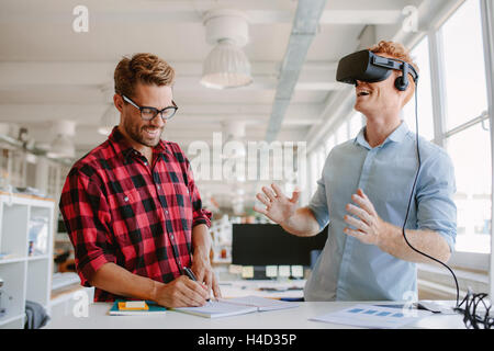 Shot of two young man standing at a table with VR goggles and writing on notepad. Developers testing virtual reality glasses in Stock Photo
