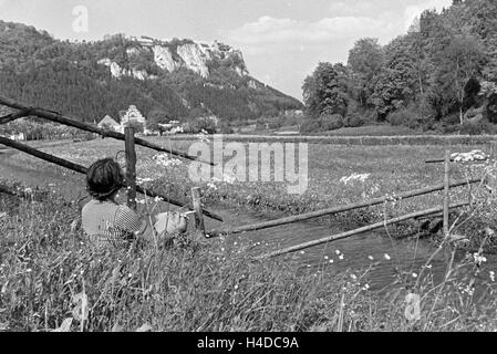 Eine junge Frau sitzt im Gras einer Wiese vor einem kleinen Bach im Schwarzwald, Deutschland 1930er Jahre. A young woman sitting in the gras at a little creek in the Black Forest, Germany 1930s. Stock Photo
