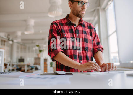 Shot of young man standing at his desk and working on computer. Businessman using computer in modern office. Stock Photo