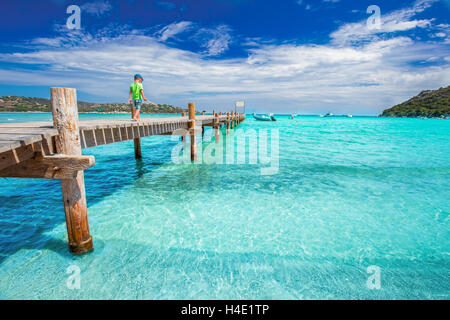 Little boy walking on the tropical bridge on Santa Giulia beach with red rocks, pine trees and azure clear water, Corsica, Franc Stock Photo