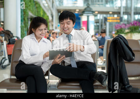 Young Asian couple business people using digital tablet pc while sitting in the airport terminal waiting for boarding. Business Stock Photo