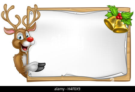 Christmas reindeer cartoon character peeking around wooden scroll sign with gold bells and holly and pointing Stock Photo