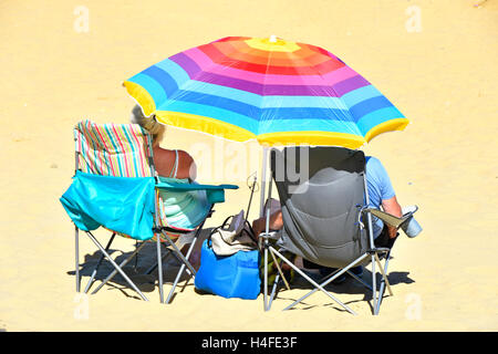 Mature couple colourful beach umbrella relaxing in fold up chairs on sandy seaside holiday beach shade from hot summer sunshine Ryde Isle of Wight UK Stock Photo