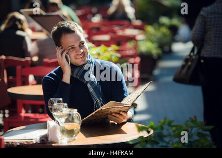 Man on cafe with smartphone talking and ordering something. Handsome young casual guy using mobile cell phone smiling happy sitting outdoors. Stock Photo