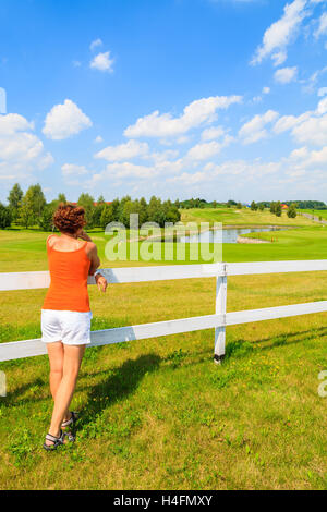 Young woman stands against a white fence and looks at golf club area on sunny summer day, Poland Stock Photo