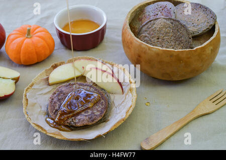 Rustic meal of buckwheat pancakes in plate and bowl made of gourds, on flower sac tablecloth, accompanied by maple syrup Stock Photo