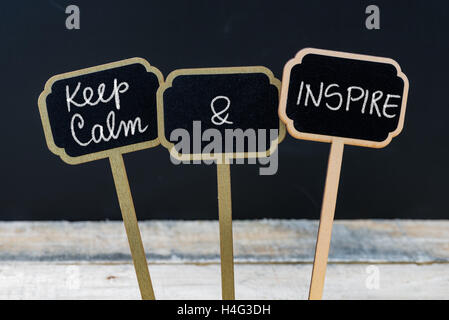Keep Calm and Inspire message written with chalk on mini blackboard labels, defocused chalkboard and wooden table in background. Fun and humor concept Stock Photo