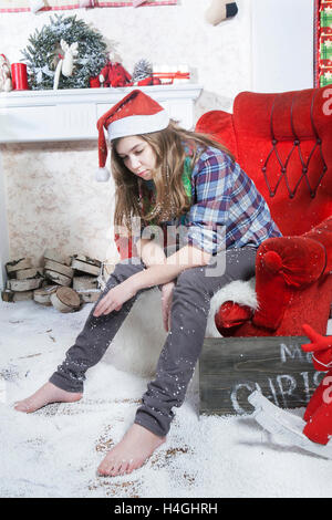 Sad girl in a plaid shirt and a cap of Santa Claus sitting on a chair. Santa Claus did not bring gifts. Stock Photo