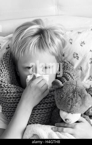 Sick child (blond boy, kid) lying in bed with teddy bear and blowing his nose Stock Photo