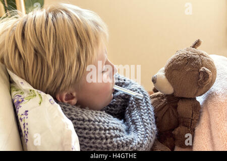 Sick child (blond boy, kid) lying in bed with thermometer and teddy bear Stock Photo