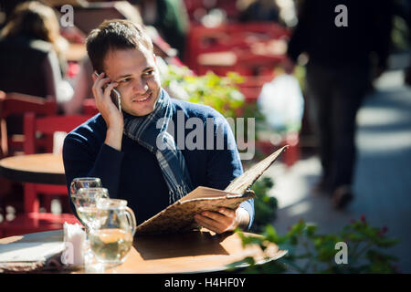 Man on cafe with smartphone talking and ordering something. Handsome young casual guy using mobile cell phone smiling happy sitting outdoors. Stock Photo