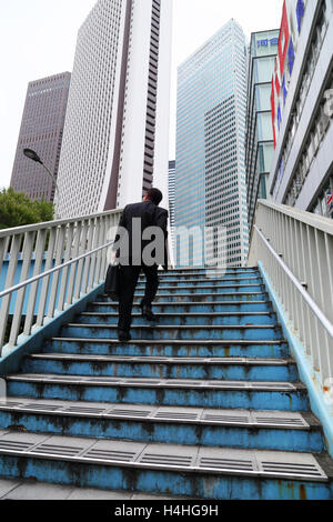 back view of a salary man in a black suit with a suitcase climbing stairs on the street with tall buildings in the background Stock Photo