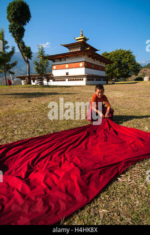 Young Buddhist monk spreads his crimson robe out to dry on the grass in front of Chimi Lhakhang near Punakha in Bhutan. Stock Photo