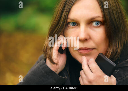 Woman with serious expression talking on mobile phone in park, dramatic light coming through treetops and falling on her face Stock Photo