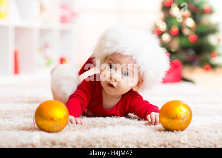 Funny baby lying on tummy wearing Santa hat and suit in front of Christmas tree Stock Photo
