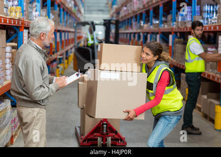 Warehouse manager noting on clipboard while female worker carrying cardboard boxes Stock Photo