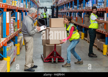Warehouse manager noting on clipboard while female worker carrying cardboard boxes Stock Photo