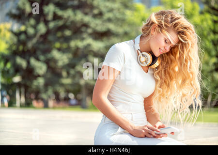 Beautiful young woman with music headphones around her neck, surfing internet on a smartphone and sitting against park backgroun Stock Photo