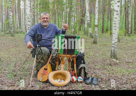 Happy senior camper is having rest in birch forest, sitting on a wicker stool and holding mandolin Stock Photo