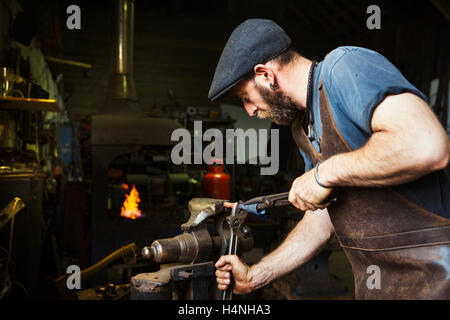 A blacksmith in a leather apron bends a cone of red hot metal in a vice using a wrench and a pair of tongs. Stock Photo