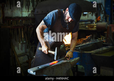 A blacksmith strikes a length of red hot metal on anvil with a hammer in a workshop. Stock Photo