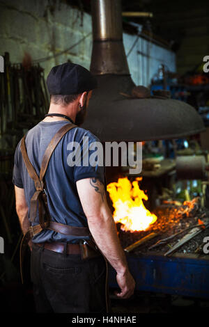 A blacksmith wearing a leather apron stands in front of a furnace in a workshop. Stock Photo