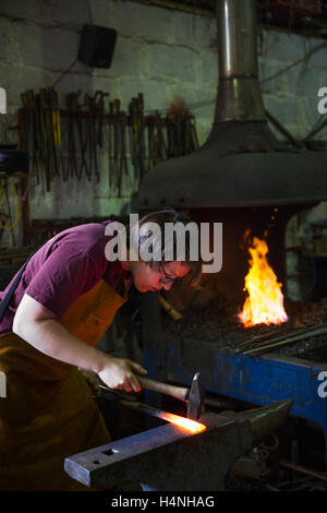 A blacksmith strikes a length of red hot metal on anvil with a hammer in a workshop. Stock Photo