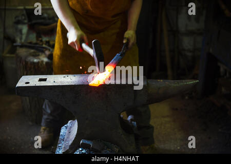 A blacksmith strikes a length of red hot metal on anvil with a hammer in a workshop. Stock Photo