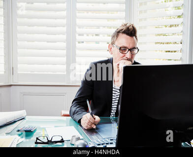 A man wearing glasses sitting at a desk in office looking at a computer screen. Stock Photo