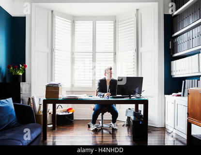 A man in suit and trainers with glasses, sitting at a desk in a bay window. A light airy office with files on shelves. Stock Photo