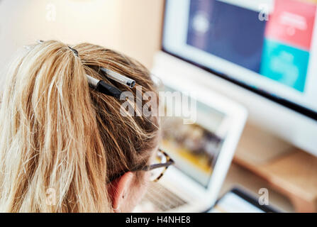 Over the shoulder view of a woman at a workstation with two pens in her hair. Laptop and computer screen graphics. Stock Photo