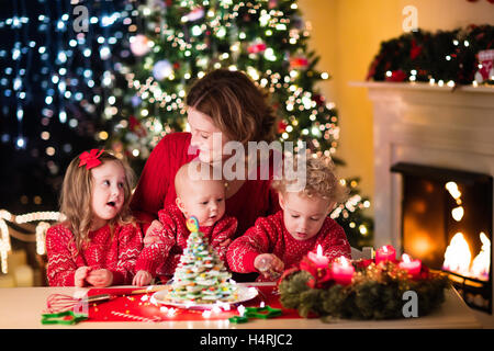 Family, mother with baby, little boy and girl making gingerbread house at fireplace in living room with Christmas tree Stock Photo