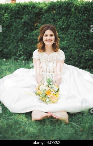 Lovely bride sitting on ground holding a bouquet smiling at camera Stock Photo