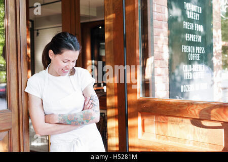 Portrait of a woman wearing a white apron with her arms crossed in a doorway, tattoos on her left arm. Stock Photo